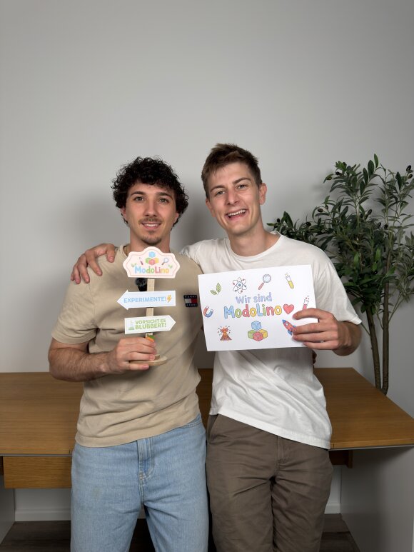 Zwei junge Männer posieren lächelnd vor heller Wand in einem schlichten Innenraum. Einer hält ein buntes ModoLino-Schild, der andere ein handgemaltes Plakat mit dem Schriftzug „Wir sind ModoLino“. Warme Farben, weiches Licht und mittige Komposition erzeugen eine freundliche, positive Stimmung.