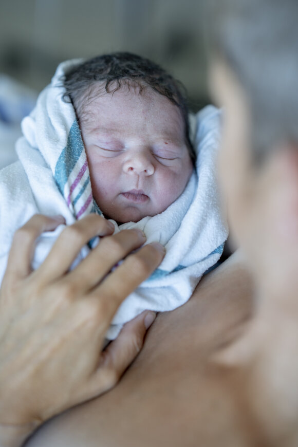 A Mother who has just given birth holds her newborn cradled in her arms as she swoons over him for the first time.  The baby is wrapped in a hospital blanket and hat to keep him warm.